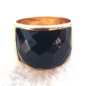 May include: A gold-coloured ring with a wide band. The band features a large, faceted, black stone. The ring is set against a white background. The ring's design is bold and eye-catching, suitable for a statement piece.