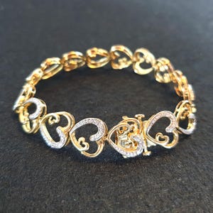 May include: Gold-coloured bracelet with heart-shaped links. Some hearts are embellished with small, sparkling stones. The bracelet has a clasp closure. The jewellery is presented on a dark surface.