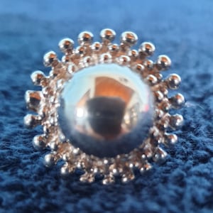 Könnte beinhalten: Nahaufnahme einer silberfarbenen, runden Brosche mit einem strukturierten, sonnenstrahlenartigen Design um eine zentrale, reflektierende Kugel. Die Brosche hat eine Vintage-Ästhetik und ist vor blauem Hintergrund platziert.