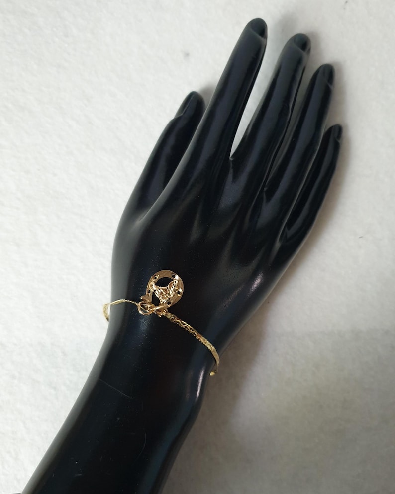 May include: A thin gold-toned bracelet featuring a textured, shimmering chain and a small, circular gold-toned charm with an embossed horseshoe design. The bracelet is delicate, with a polished finish that reflects light across its surface.