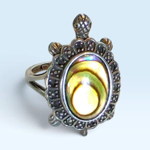 May include: A silver-toned ring featuring a turtle-shaped centerpiece. The turtle shell is inlaid with an iridescent abalone shell oval, surrounded by a textured metal border with small bead-like details. The ring band is split at the sides.