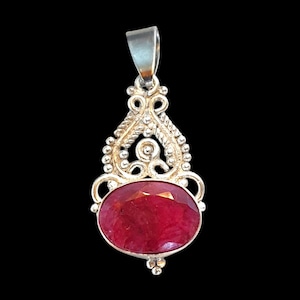 May include: A silver pendant featuring an oval-shaped, faceted deep red gemstone set in a decorative metal frame with intricate scrollwork, beaded accents, and a smooth bail at the top for attachment to a chain or cord necklace.