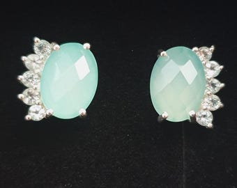 May include: A pair of silver-toned stud earrings. Each earring features a large, light blue, oval-shaped gemstone with a faceted cut. Small, clear gemstones are clustered around the oval stone, creating a decorative accent. The background is black.