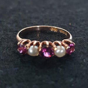 Peut inclure: Une bague dorée ornée de pierres précieuses et de perles alternées. La bague comporte cinq pierres : deux perles et trois pierres précieuses magenta. La bague est posée sur un fond sombre.