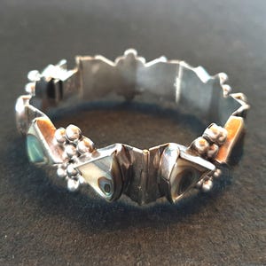 May include: A silver-coloured hinged bracelet with geometric shapes and iridescent abalone shell inlays. The bracelet features small, round, silver-coloured accents and a textured, angular design. The bracelet is shown on a dark surface.