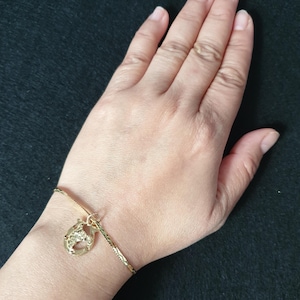 May include: A gold-toned chain bracelet featuring a textured, horseshoe-shaped charm. The bracelet has a delicate, link-style chain with a reflective finish, and the charm is attached by a small circular jump ring, creating a simple accessory.