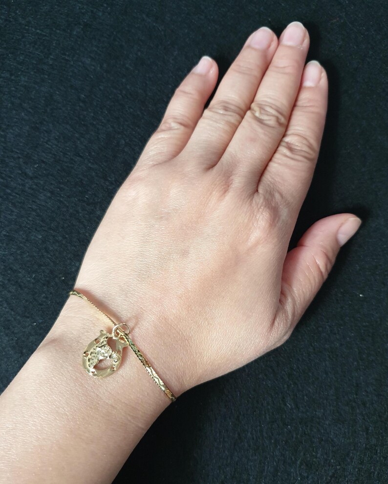 May include: A gold-toned chain bracelet featuring a textured, horseshoe-shaped charm. The bracelet has a delicate, link-style chain with a reflective finish, and the charm is attached by a small circular jump ring, creating a simple accessory.