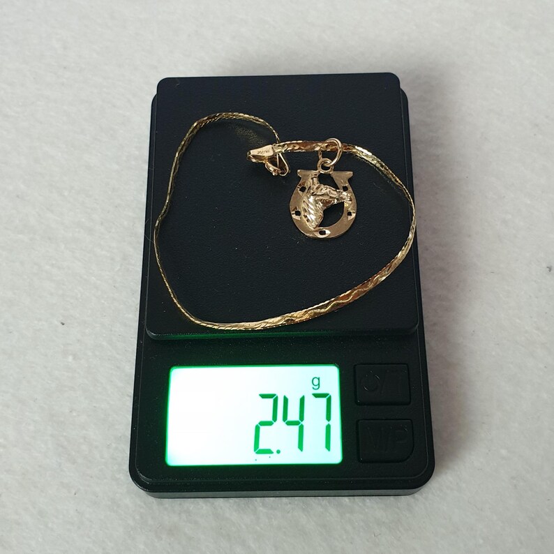 May include: A gold-coloured chain necklace featuring a horseshoe pendant with a horse head design. The jewellery is placed on a digital scale that displays a weight of 2.47 grams, providing a clear measurement of the item's mass in a professional setting.