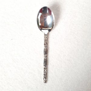 Peut inclure: Une cuillère en métal argenté dotée d'un cuilleron ovale lisse et très réfléchissant, ainsi qu'un long manche fin orné d'une série de figures sculptées répétitives et empilées verticalement, s'étendant du col jusqu'à la base du manche.