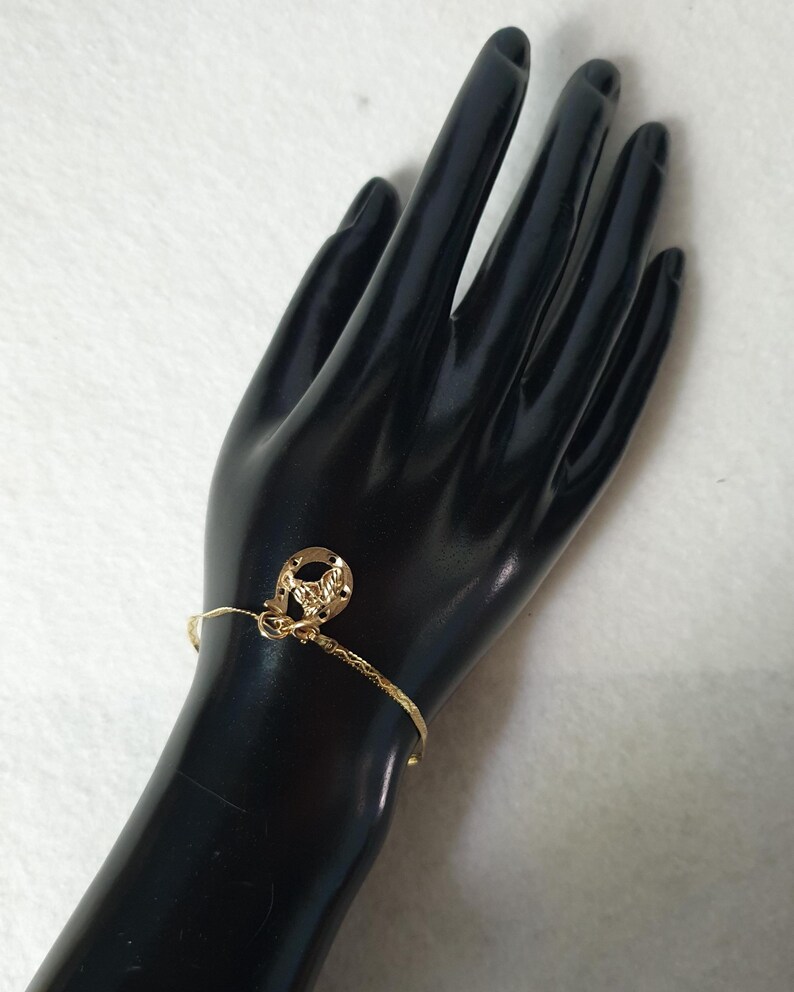 May include: A gold-toned bracelet featuring a textured, thin band and a single charm shaped like a horseshoe with a central horse head design. The metal charm is attached to the bracelet with a small jump ring, creating a simple, metallic accessory.