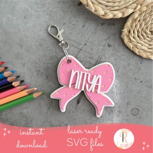 May include: A pink bow-shaped keyring with the name "NIYA" in white lettering. The bow has a floral pattern and a silver clasp. The image also includes coloured pencils and text that says "instant download" and "laser ready SVG files."