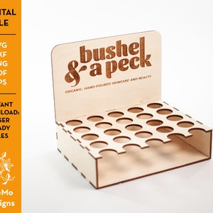 Puede incluir: Un expositor de madera con un diseño de corte para sostener botellas pequeñas. El expositor tiene un letrero que dice "bushel & a peck organic, hand-poured skincare and beauty".