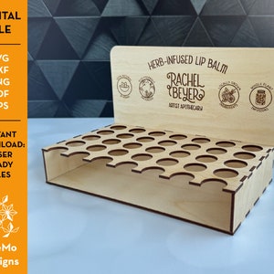 Puede incluir: Un expositor de madera para bálsamo labial con 24 ranuras. El expositor está cortado con láser y grabado con el texto "Bálsamo labial con infusión de hierbas", "Rachel Beyer", "Artist Apothecary", "Ingredientes cultivados localmente" y "Infusiones de plantas enteras".
