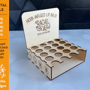 May include: A laser-cut wooden display stand for lip balm. The stand has multiple circular openings to hold lip balm tubes. The top of the stand features the text "HERB-INFUSED LIP BALM" and "RACHEL BEYER ARTIST APOTHECARY". The stand is a digital file.