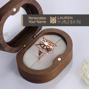 May include: A rose gold ring with a geometric design and a blue gemstone. The ring is in a wooden box with the words "Personalize Your Name" and "LAUREN" on the lid.