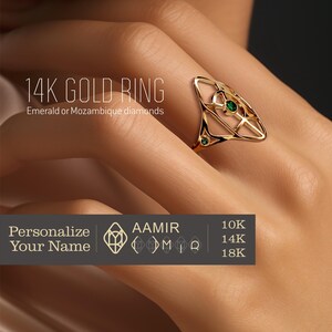 Könnte beinhalten: Ein 14 Karat Goldring mit geometrischem Design und zwei kleinen grünen Edelsteinen. Der Ring befindet sich an einem Finger eines Models.