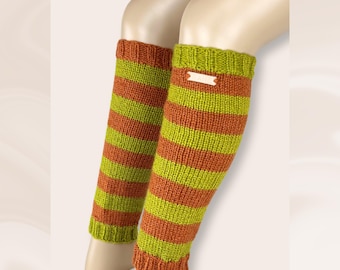 May include: A pair of leg warmers featuring horizontal stripes in lime green and burnt orange. The knit fabric has ribbed cuffs at the top and bottom. A small rectangular wooden tag is attached to the upper section. Text reads: CRAFTED. NOT PRODUCED. Handknitted for you.