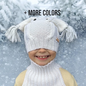 May include: White knitted hat with ear flaps and a pom-pom on each side. The hat is pulled down over the child's eyes, and they are smiling. The text '+ MORE COLORS' is visible at the top of the image.