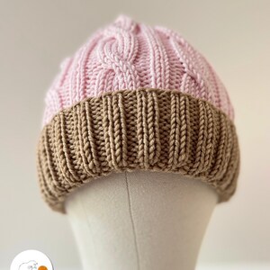 Op de afbeelding: Een tweekleurige gebreide beanie-muts. Het bovenste gedeelte is lichtroze met een kabelbreipatroon, terwijl de rand een effen bruine kleur heeft. De hoed wordt op een mannequinhoofd getoond.