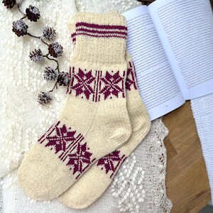 May include: Cream-coloured knitted socks with a burgundy geometric pattern. The socks have a ribbed cuff with burgundy stripes. The socks are displayed on a white lace surface, with a book and decorative pinecones in the background.