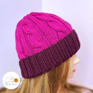 Op de afbeelding: Een gebreide beanie in een levendige fuchsia kleur met een diepe bordeauxrode geribbelde rand. De hoed heeft een kabelgebreid ontwerp op de kroon. Een winteraccessoire.