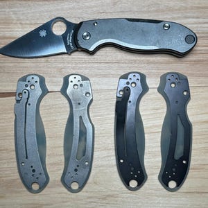 May include: Five different knife scales, two are silver, two are black, and one is gray. The scales are made of metal and have a textured surface. The scales are for a folding knife.