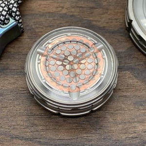 May include: A silver fidget spinner with a copper center featuring a honeycomb pattern. The spinner has a textured outer ring.