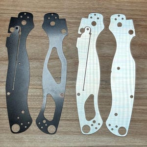 May include: Four metal knife scales, two are black and two are silver. The scales are for a folding knife and have cutouts for the blade and handle.