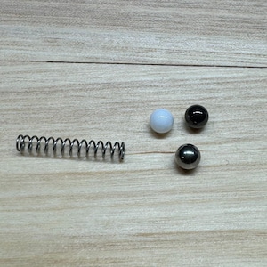 May include: A metallic helical compression spring positioned horizontally next to three small spherical balls. The balls consist of one solid white sphere, one solid black sphere, and one polished metallic silver sphere, all arranged in a triangular formation.