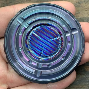 Op de afbeelding: Een rond metalen fidget-speelgoed met een blauw, paars en gouden swirl-ontwerp in het midden. De buitenring heeft een getextureerde, geborstelde afwerking.