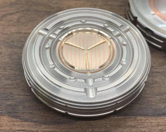 May include: A round, silver-colored watch face with a concentric circle design. The watch face has a smaller, circular, gold-colored dial with a white and pink striped pattern and three gold-colored hands.