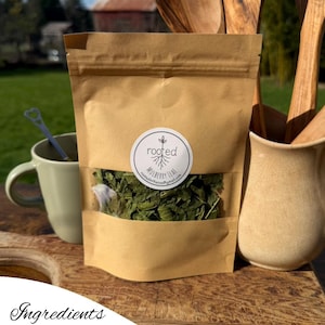 May include: A brown paper pouch with a clear window containing dried green mulberry leaves. A circular white label on the front features a tree root graphic and the text rooted MULBERRY LEAF. The pouch is positioned near wooden utensils.