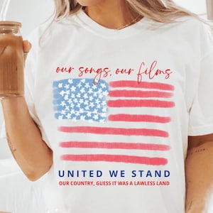 Op de afbeelding: Wit T-shirt met een aquarelontwerp van de Amerikaanse vlag en de tekst "our songs, our films" erboven en "UNITED WE STAND" eronder. Het shirt bevat ook de zin "OUR COUNTRY, GUESS IT WAS A LAWLESS LAND".