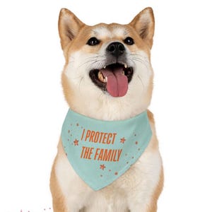 Puede incluir: Un perro Shiba Inu con un pañuelo azul claro con el texto "I PROTECT THE FAMILY" en rojo. El perro está sentado y mirando hacia arriba con la lengua fuera. El fondo es blanco.