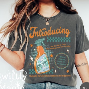 Pode incluir: Camiseta cinza escuro com a palavra "Introducing" em escrita laranja acima de um gráfico de um borrifador laranja rotulado "Opalite". A camiseta inclui o texto "Just One Spray Turns Your Problems Into Your Paradise."