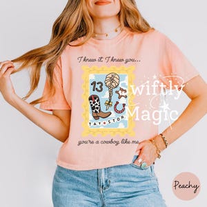 May include: A peach-colored short-sleeve t-shirt featuring a graphic design of a postage stamp with the text I knew it, I knew you... and you're a cowboy like me. The stamp includes a cow print boot, a braid, a horseshoe, and the number 13.