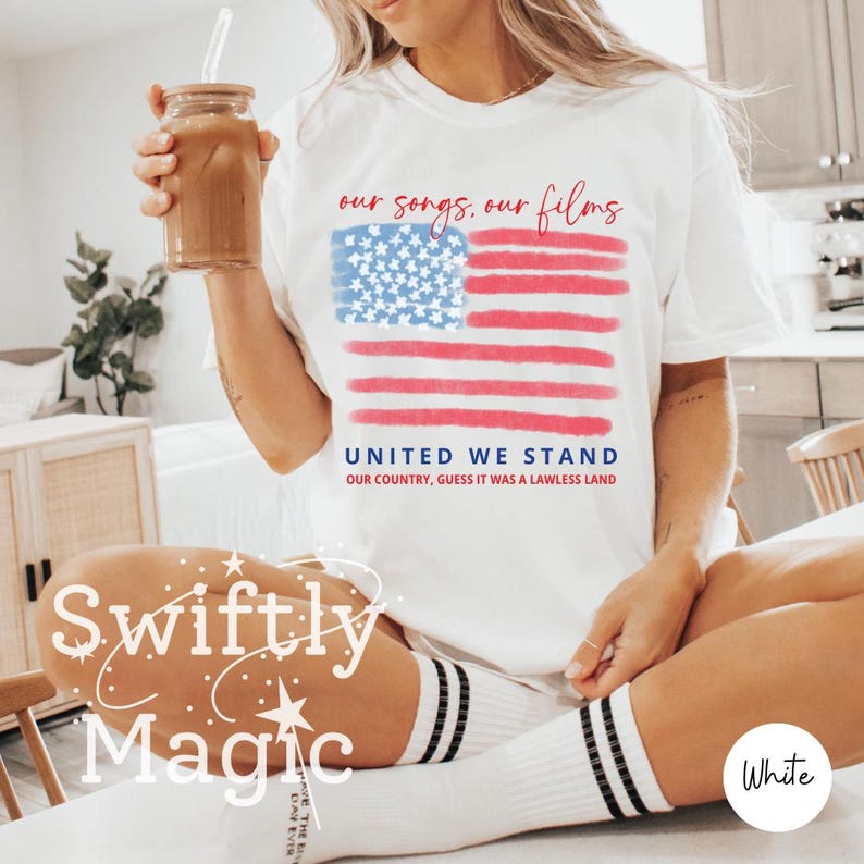Op de afbeelding: Wit T-shirt met een aquarelontwerp van de Amerikaanse vlag en de tekst "our songs, our films" erboven en "UNITED WE STAND" eronder. Het shirt bevat ook de zin "OUR COUNTRY, GUESS IT WAS A LAWLESS LAND".