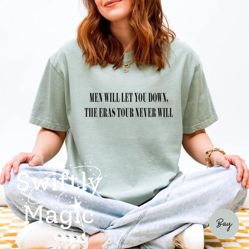 May include: Sage green t-shirt with the text "MEN WILL LET YOU DOWN. THE ERAS TOUR NEVER WILL" in black. The shirt is a relaxed fit and is paired with light wash jeans. The image is taken in natural light.