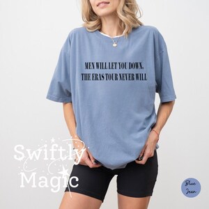 May include: A light blue t-shirt with the text "MEN WILL LET YOU DOWN. THE ERAS TOUR NEVER WILL" in black. The shirt is a relaxed fit and has short sleeves. The image also includes black shorts.