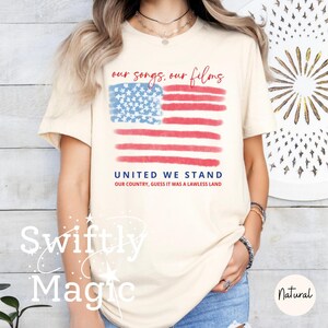 Op de afbeelding: Cr&egrave;mekleurig T-shirt met een gestileerd Amerikaans vlagontwerp in rood en blauw. De tekst "our songs, our films" staat boven de vlag en "UNITED WE STAND" eronder. Extra tekst luidt "OUR COUNTRY, GUESS IT WAS A LAWLESS LAND".