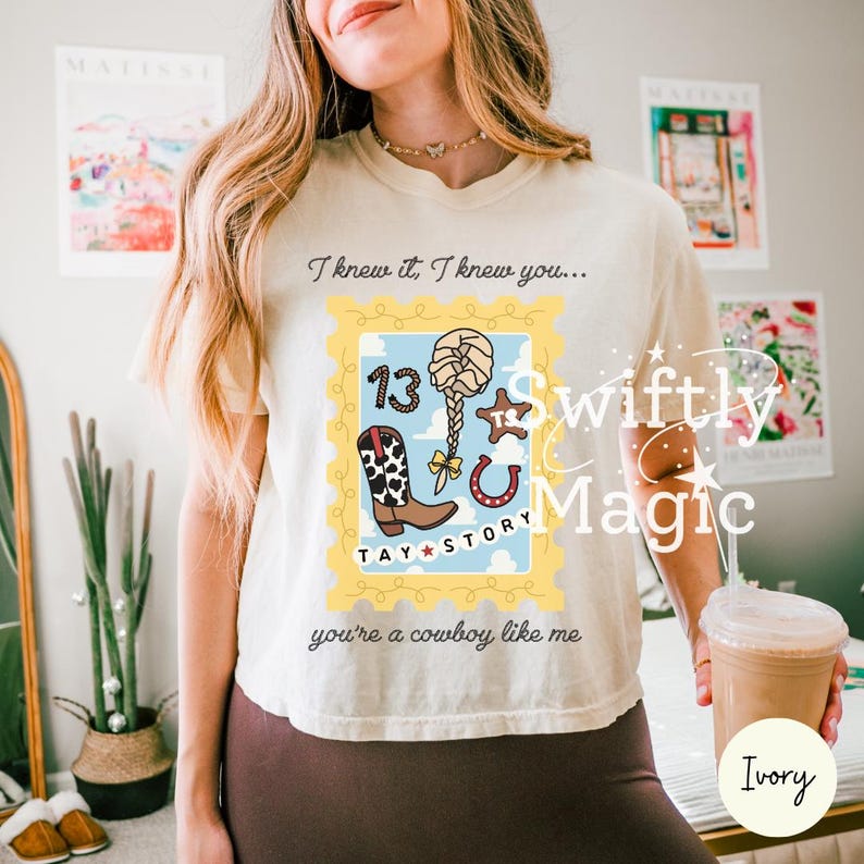 May include: An ivory t-shirt featuring a graphic print of a yellow postage stamp frame containing a cowboy boot, a braided hairstyle, a star, and a horseshoe. The text reads: I knew it, I knew you... TAY STORY you're a cowboy like me.