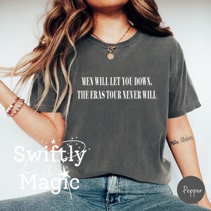 May include: Dark gray t-shirt with the text "MEN WILL LET YOU DOWN. THE ERAS TOUR NEVER WILL" in white. The shirt is a casual style, perfect for everyday wear. The word "Swiftly Magic" is also visible.