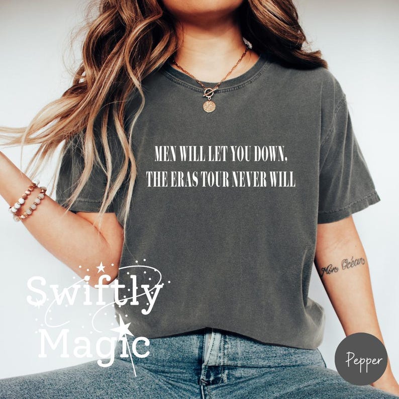 May include: Dark gray t-shirt with the text "MEN WILL LET YOU DOWN. THE ERAS TOUR NEVER WILL" in white. The shirt is a casual style, perfect for everyday wear. The word "Swiftly Magic" is also visible.