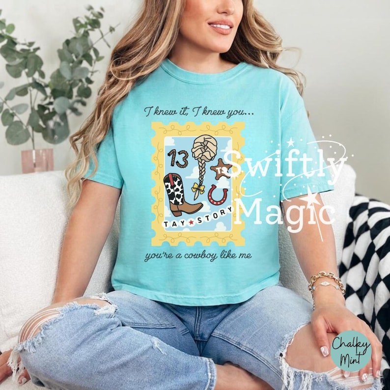 May include: A light blue t-shirt featuring a yellow postage stamp graphic with the text I knew it, I knew you... and you're a cowboy like me. The graphic includes the number 13, a braided ponytail, a cow print boot, a horseshoe, and Tay Story.