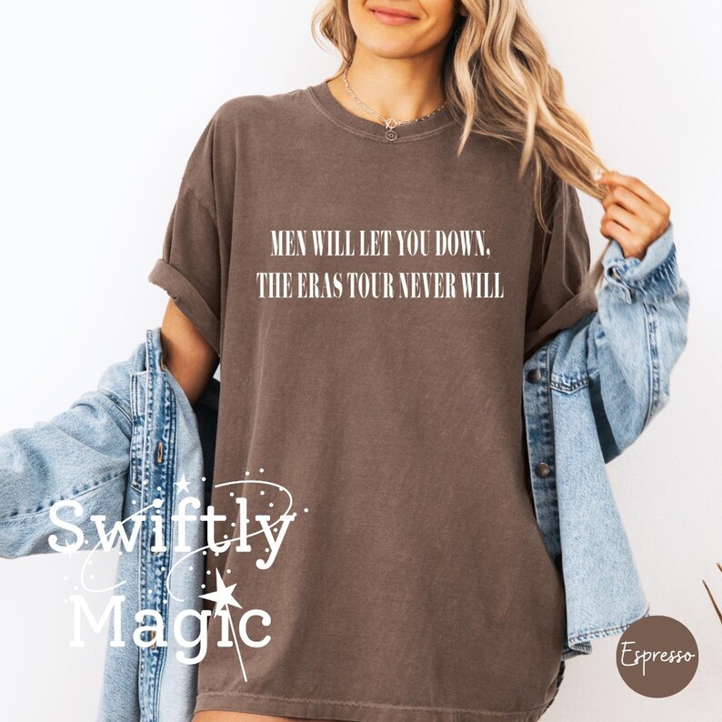May include: A brown t-shirt with the text "MEN WILL LET YOU DOWN. THE ERAS TOUR NEVER WILL" in white. The shirt is part of a clothing collection. The brand name "Swiftly Magic" is visible in the lower left corner.