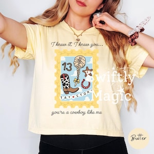 May include: A pale yellow t-shirt featuring a graphic design shaped like a postage stamp. The print includes a cowboy boot, a braided hairstyle, a horseshoe, and the number 13. Text reads: I knew it, I knew you... TAY STORY you're a cowboy like me.