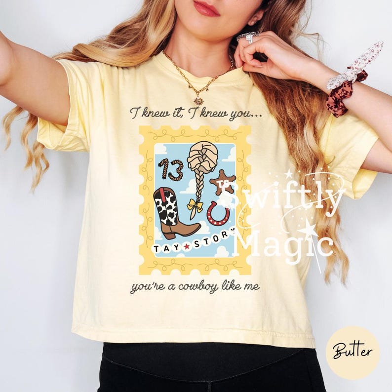 May include: A pale yellow t-shirt featuring a graphic design shaped like a postage stamp. The print includes a cowboy boot, a braided hairstyle, a horseshoe, and the number 13. Text reads: I knew it, I knew you... TAY STORY you're a cowboy like me.
