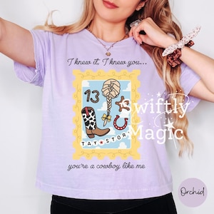 May include: A light purple t-shirt featuring a graphic design shaped like a postage stamp. The design includes a cowboy boot, a braided hairstyle, a horseshoe, and the number 13. Text reads: I knew it, I knew you... TAY STORY you're a cowboy like me.
