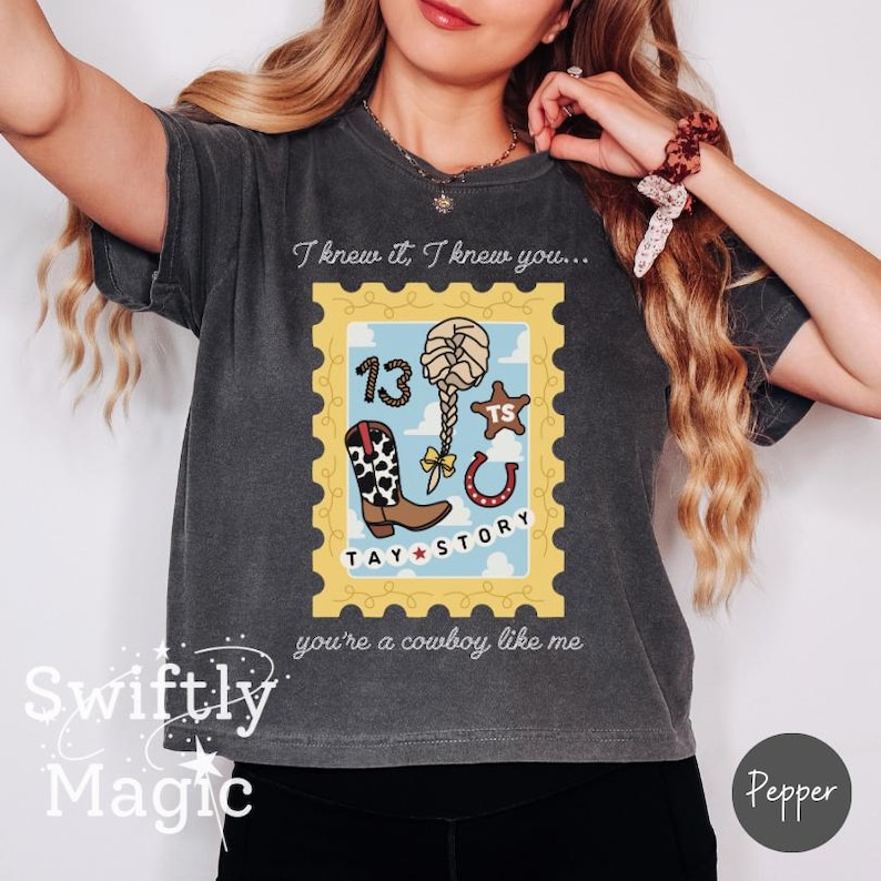 May include: A charcoal gray t-shirt featuring a graphic of a yellow postage stamp frame containing a cow print boot, a braided hairstyle, a horseshoe, a star with the letters TS, and the number 13. The text reads I knew it, I knew you... TAY STORY you're a cowboy like me.