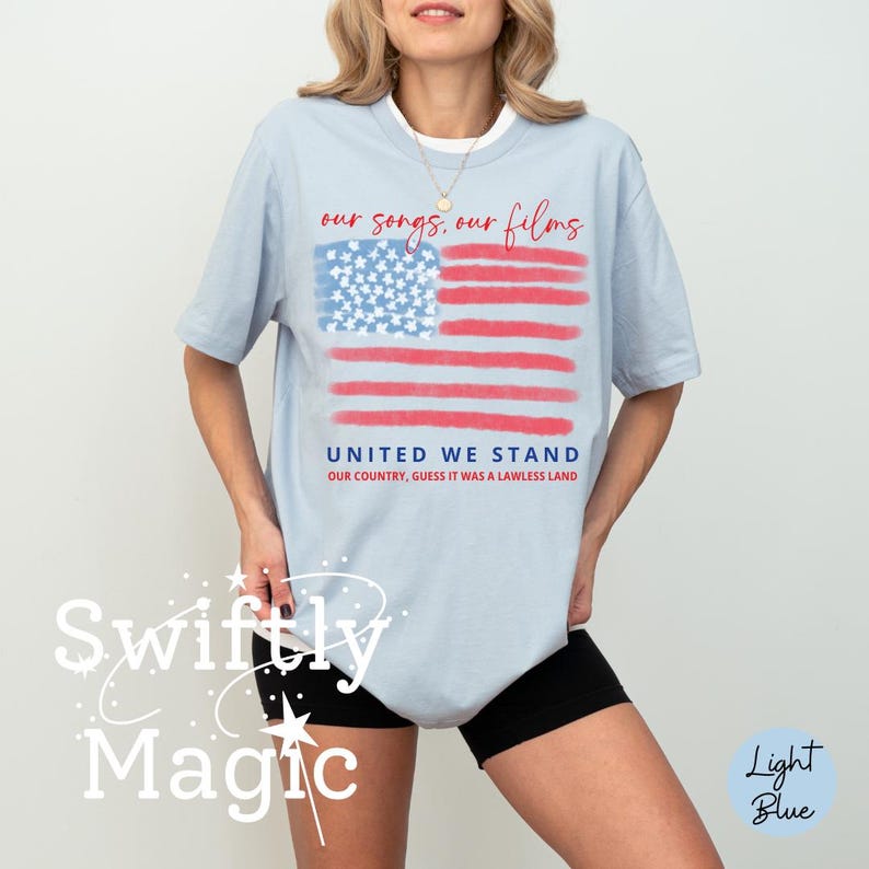 Op de afbeelding: Lichtblauw T-shirt met een gestileerd ontwerp van de Amerikaanse vlag. De vlag heeft rode strepen en een blauw sterrenveld. De tekst op het shirt luidt "our songs, our films" en "UNITED WE STAND".